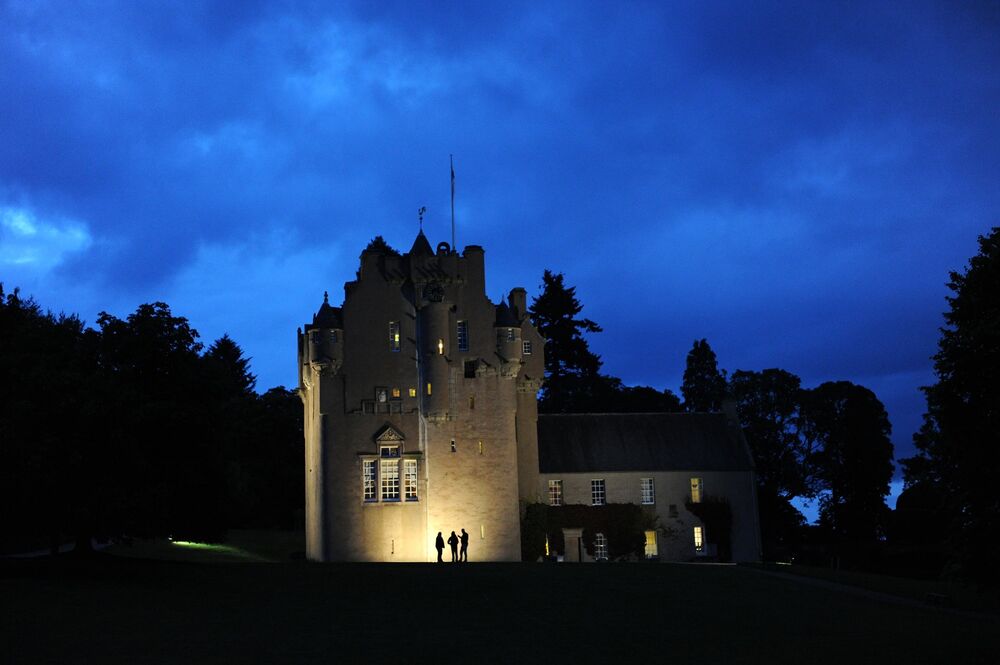 Crathes Castle at night, with a very faint light left in the sky. There are floor lights at ground level, lighting up part of the exterior. Three silhouetted figures are caught in that light.