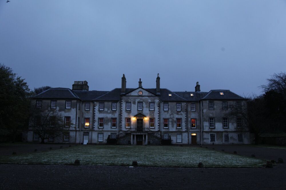 A view of Newhailes House at dusk, with very little daylight remaining. Two windows in the centre of the house and one round window in the attic are lit from inside. Frost lies on the ground in front of the house.