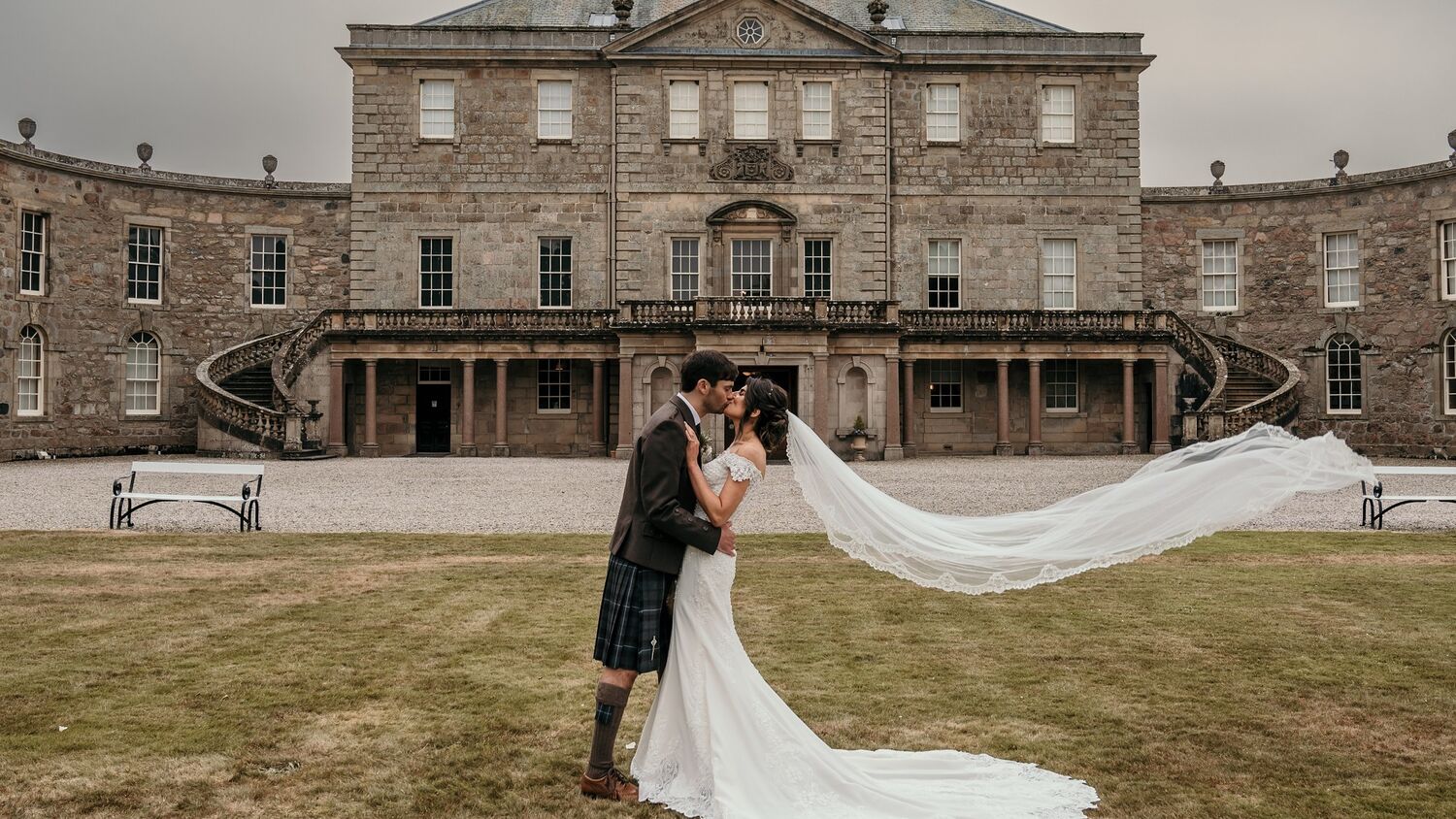A bride and groom kiss on the lawn in front of Haddo House. The wind is blowing the bride's long veil right out behind her.
