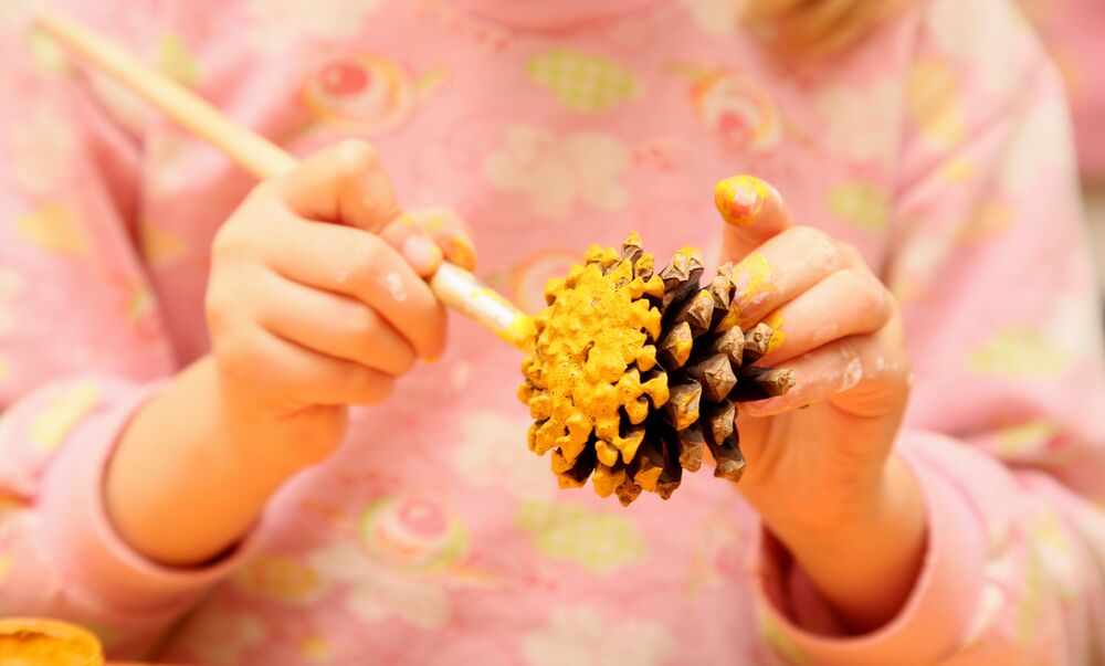 A young girl holds a pinecone in one hand and paints the bottom with yellow paint.