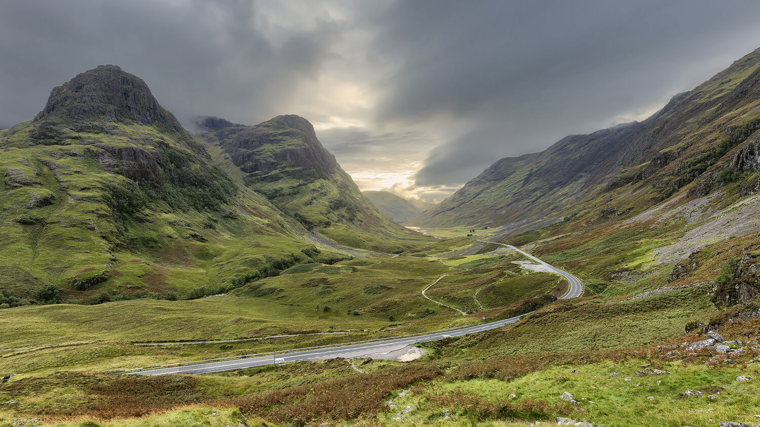 A view of Glencoe, with the golden sun shining through the dark clouds at the far end. The road snakes through the bottom of the glen, as towering mountains rise either side.