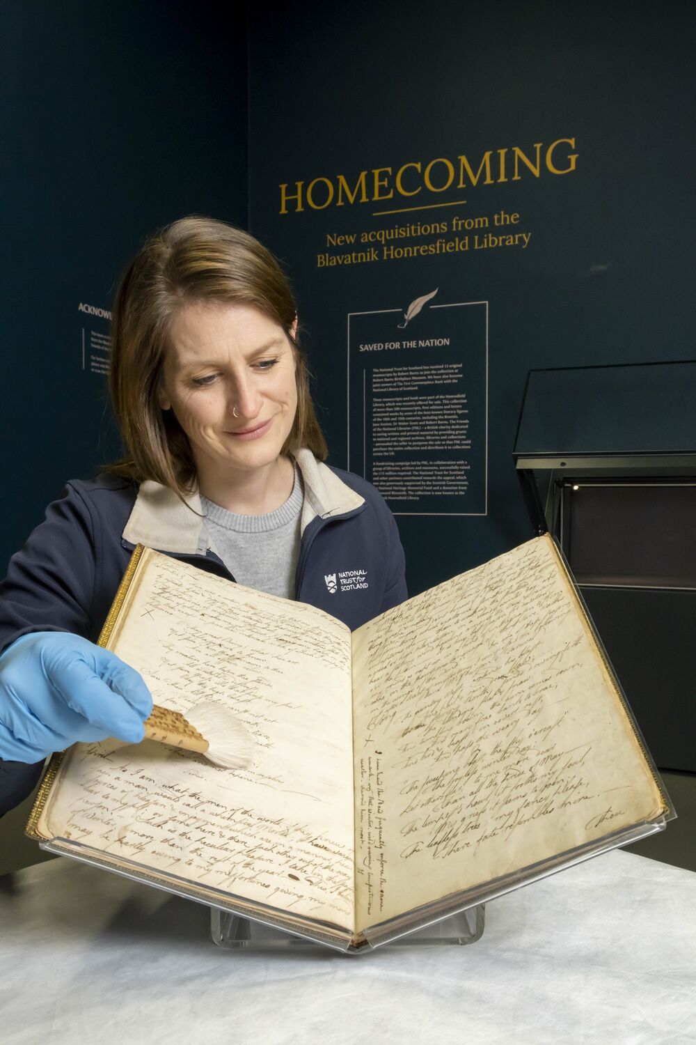 A smiling lady holds open a large book, filled with Burns's handwritten notes, and shows it to the camera. She holds a small brush in one hand and wears blue nitrile gloves.
