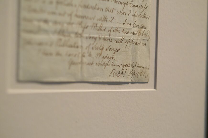 A close-up photo of the end of a handwritten letter from Robert Burns, where he has signed his name on the bottom right corner. Burns has used a brownish ink on parchment-style paper. The letter has been mounted on cream card.