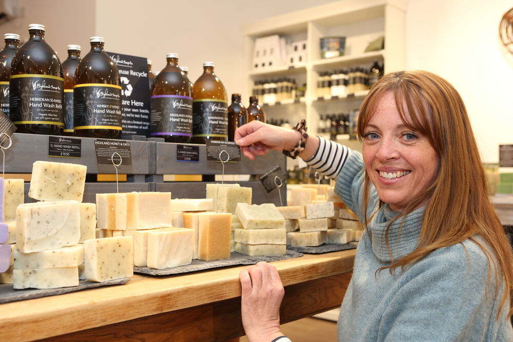 A woman in a light grey jumper with long red hair crouches down by a display table filled with soap bars and bottles.