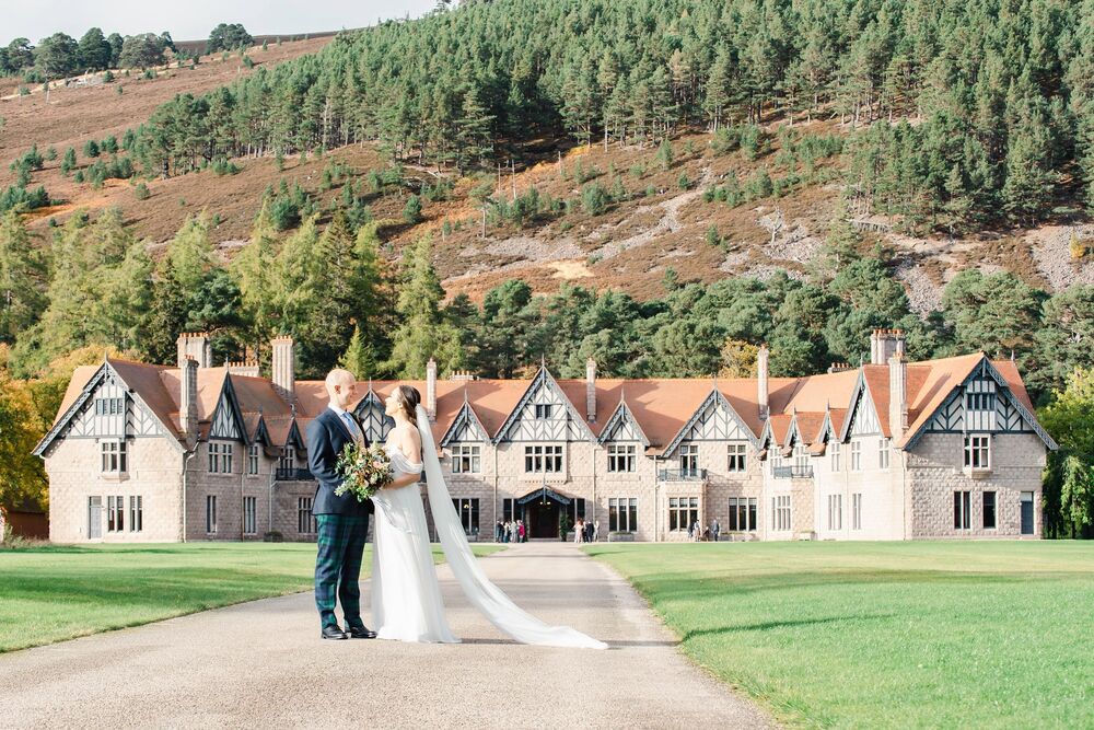 A bride and groom stand on a wide path across a lawn, with the magnificent Mar Lodge behind them.