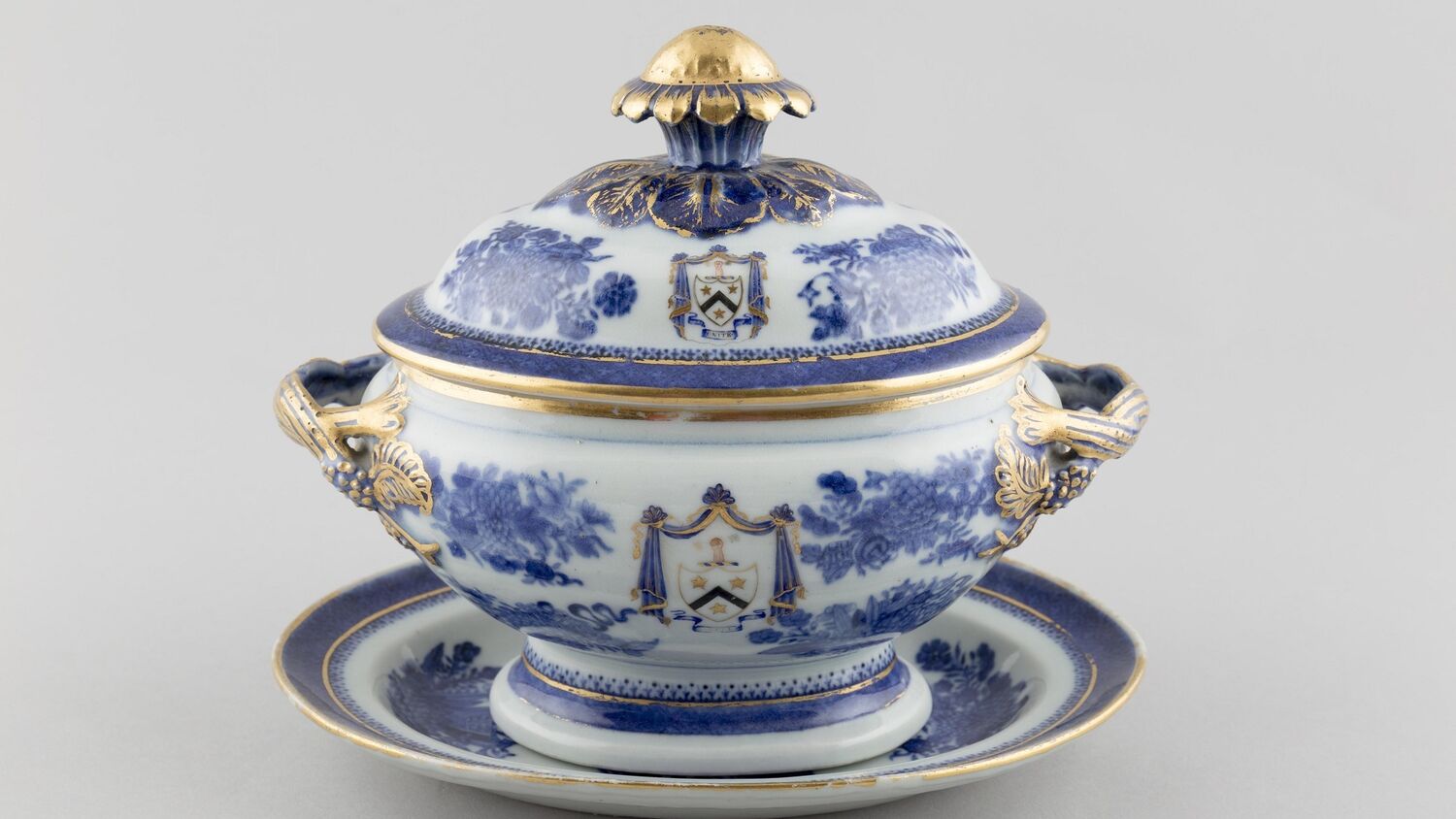 An elaborate porcelain pot with a lid stands on a little tray, against a plain grey background. It is decorated with a blue and gold pattern and has a family crest at the centre.