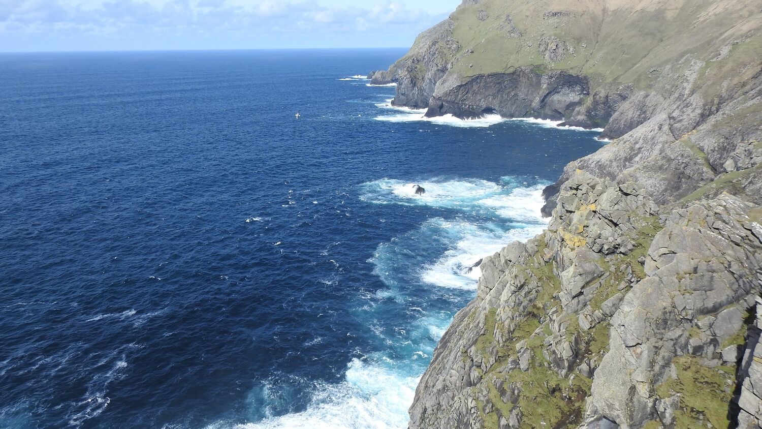 A view of the rocky cliffs of St Kilda. The deep blue sea on the left is crashing into the rocks, with a wide strip of white breakers. A seabird wheels in the air close to shore.