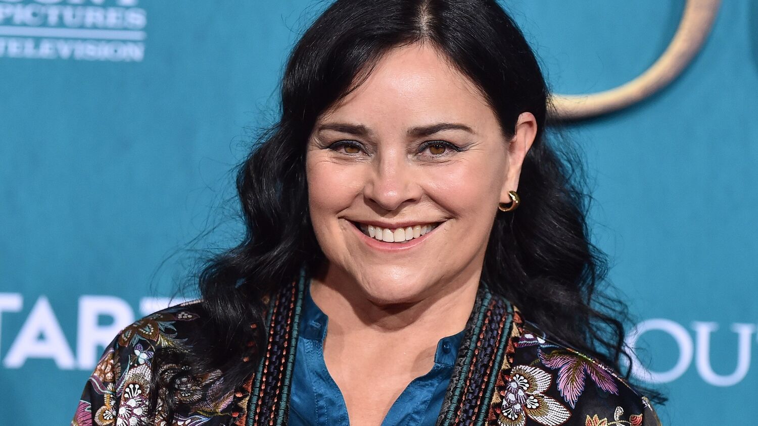 A close-up photo of Diana Gabaldon standing in front of an awards screen at a ceremony. She has long dark hair and is smiling at the camera.
