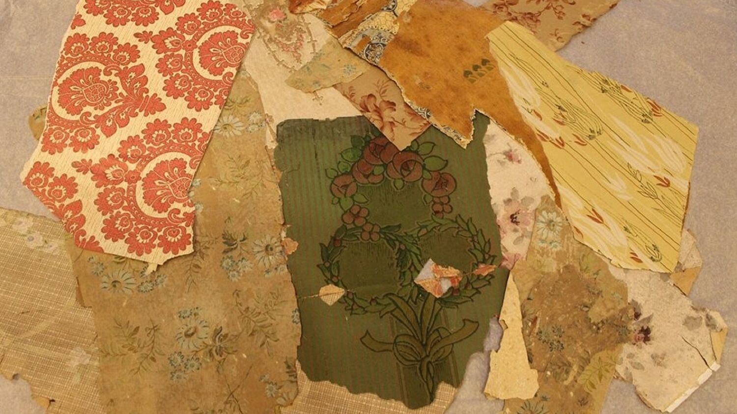 Scraps of old wallpaper are laid upon a table. Most have some sort of floral pattern and are a deep red or dark green in colour.