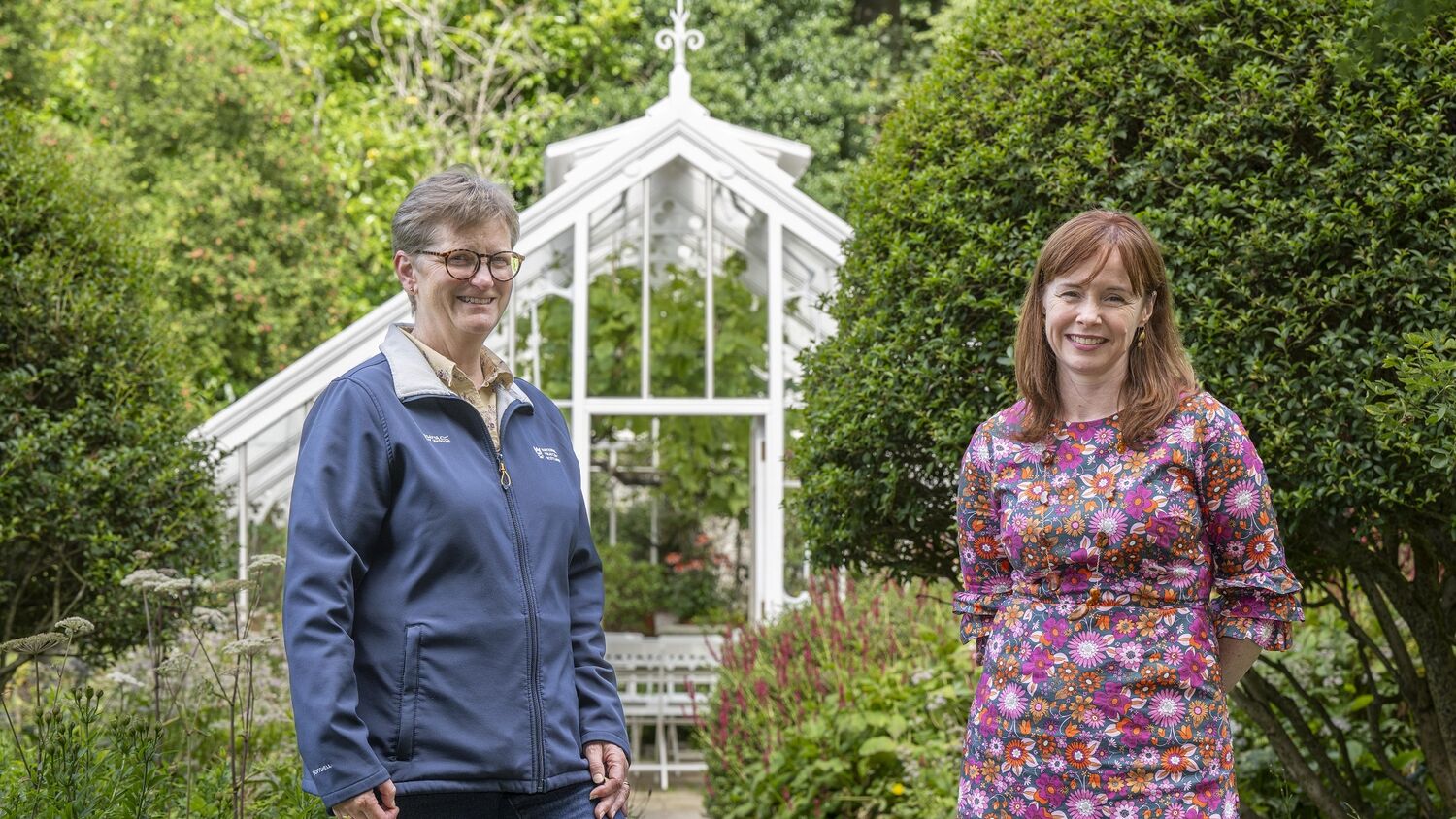 Two women stand in a garden, with an Edwardian glasshouse behind them. The lady on the left wears a navy National Trust for Scotland jacket. The lady on the right wears a floral dress.