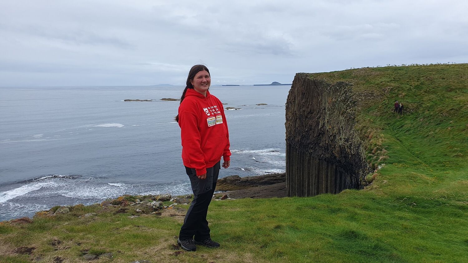 A young woman stands on a grassy clifftop, with the sea beneath her. She wears a bright red t-shirt.