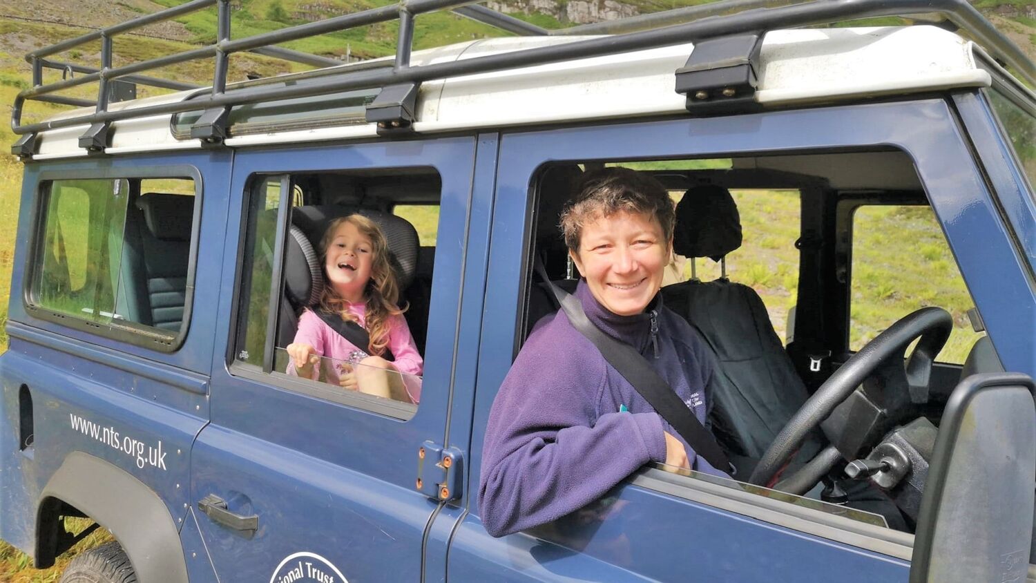 A National Trust for Scotland ranger sits in the driver's seat of a Land Rover, and leans through the open window. A young girl sits in a car seat in the back, smiling.