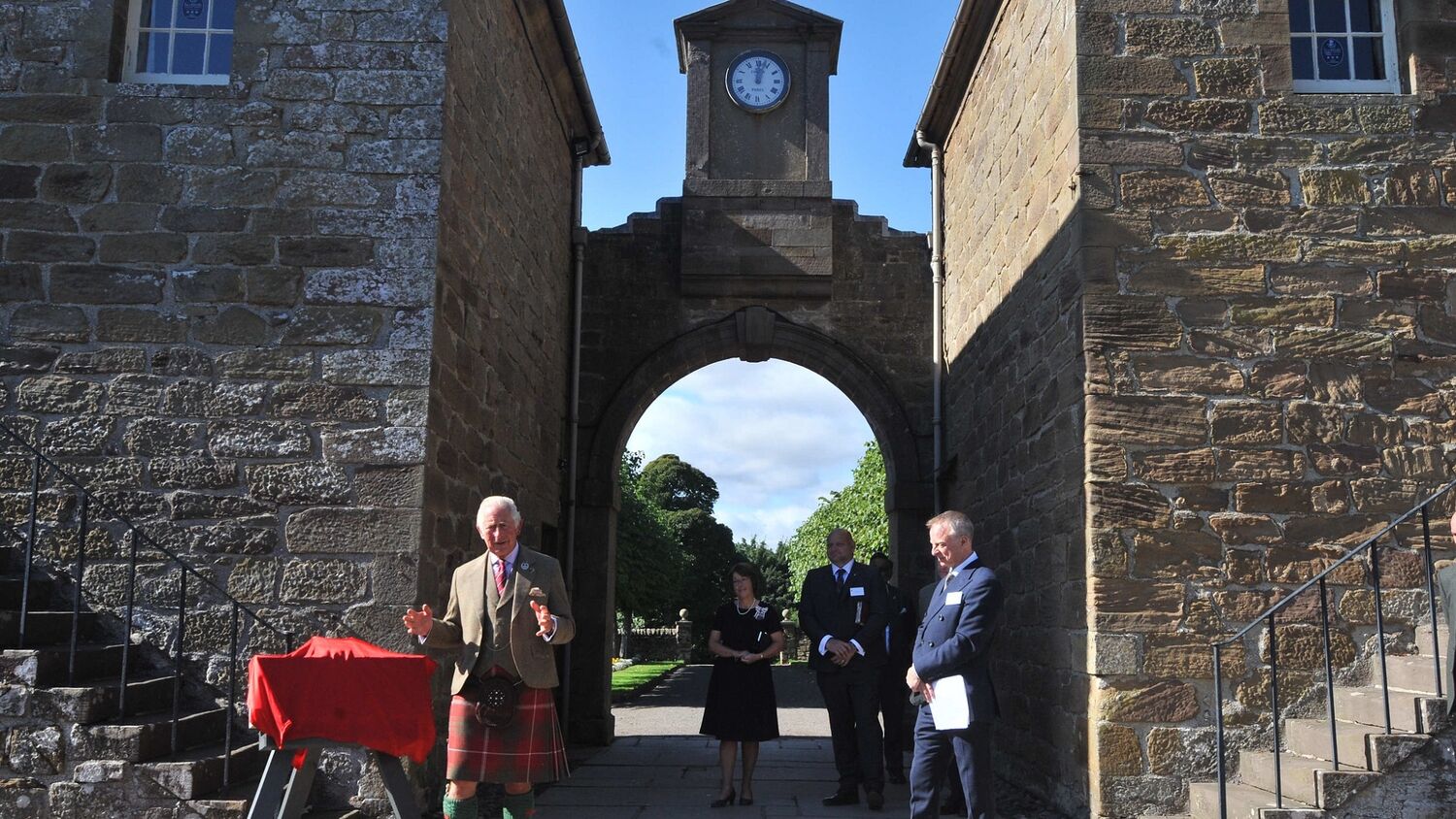 Prince Charles and some other smartly dressed officials stand in a courtyard beside some stone steps. Prince Charles stands beside a plaque covered by a red sheet.