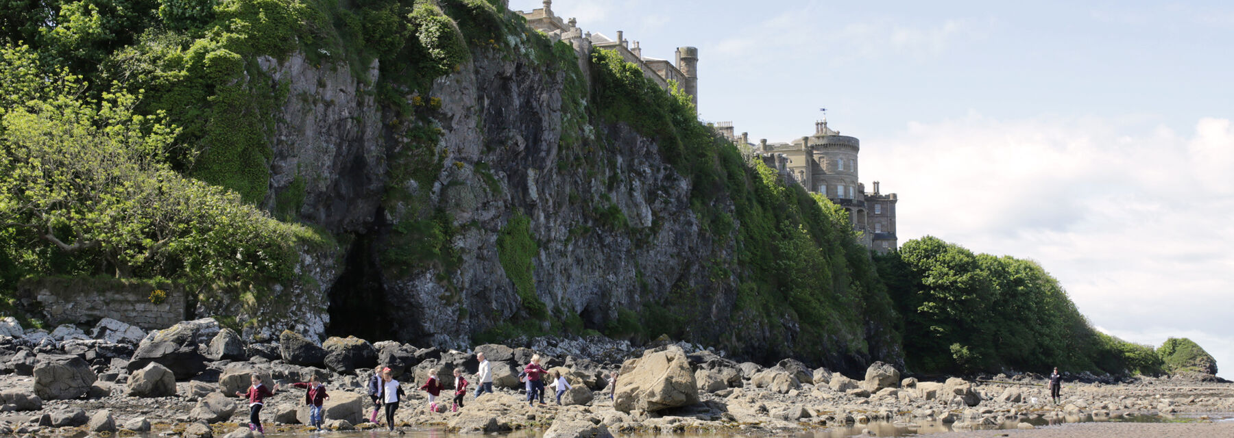 A view looking up a cliff to a grand castle on the top. Rock pools on the sandy beach are in the foreground. Boulders lie strewn on the beach beneath the cliff.