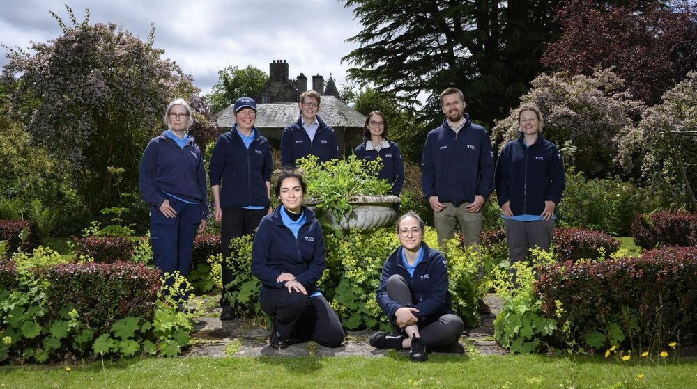 8 people stand or sit around a stone urn filled with plants in a garden. A large house is in the background. There are 6 women and 2 men. All wear navy National Trust for Scotland fleeces.