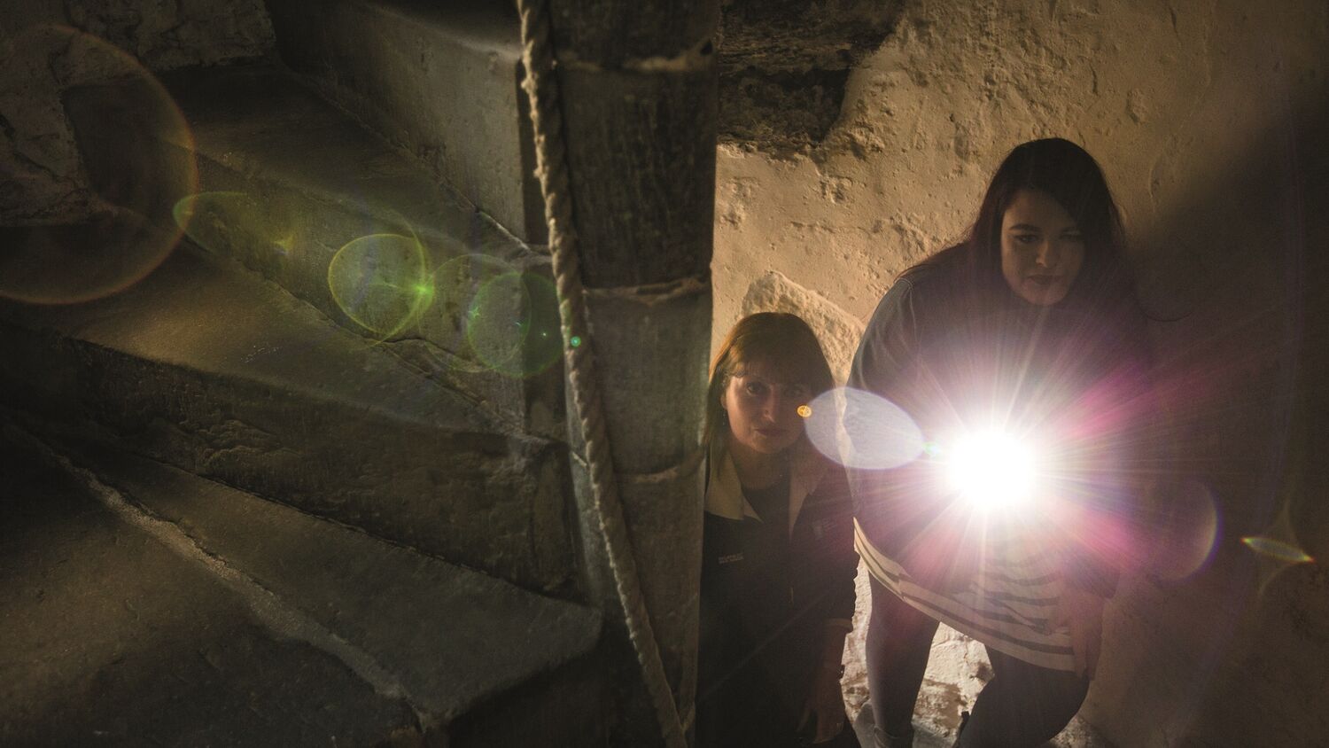 Two women walk up a narrow stone spiral stairwell, looking anxious. The lady in front carries a bright torch to light the stairs above.