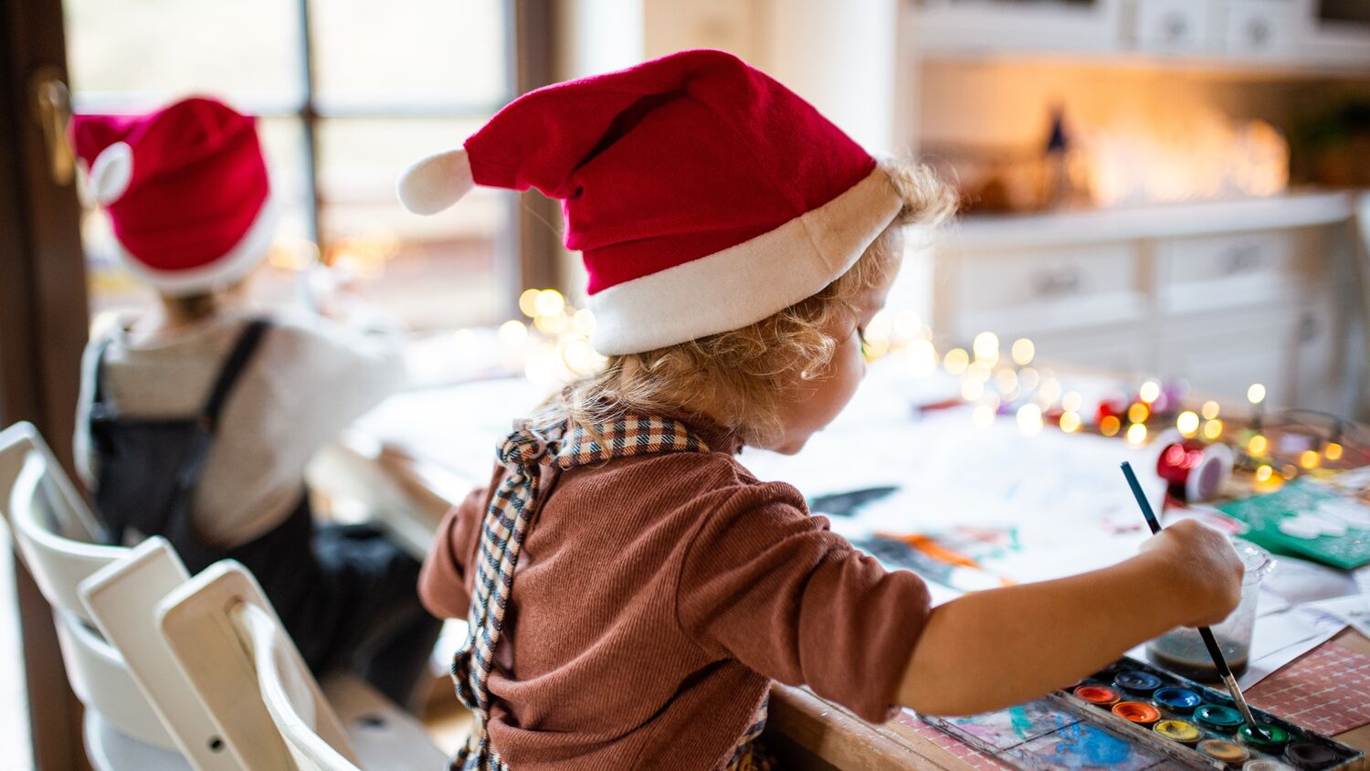 Two young children wearing Santa hats sit at a low table, with their backs to the camera. The child nearest the camera is painting.
