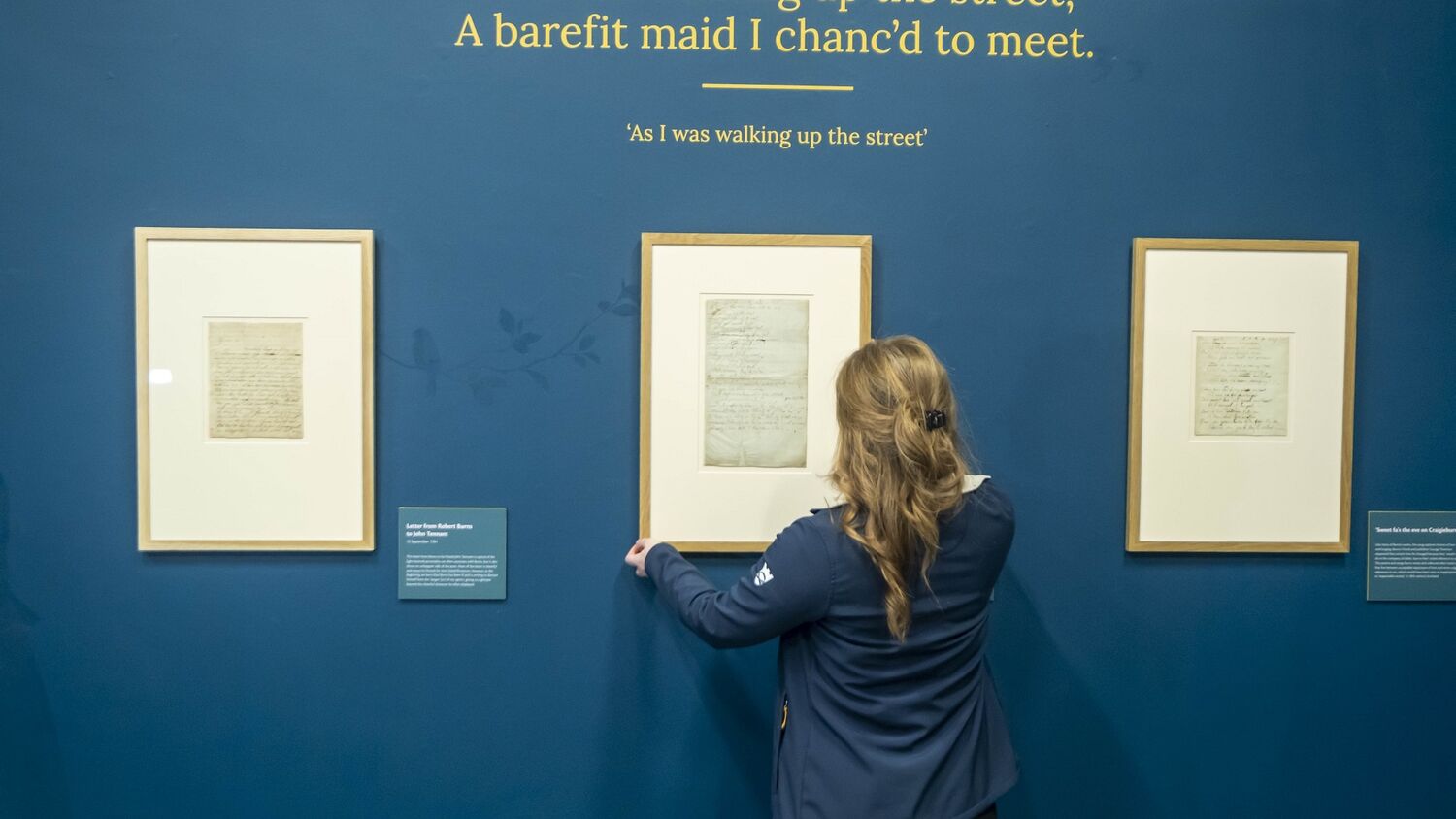 A lady stands with her back to the camera, adjusting a framed manuscript hanging on a blue wall. Other framed manuscripts hang beside it as well as interpretation panels. A Burns quote is written on the wall above the manuscripts.