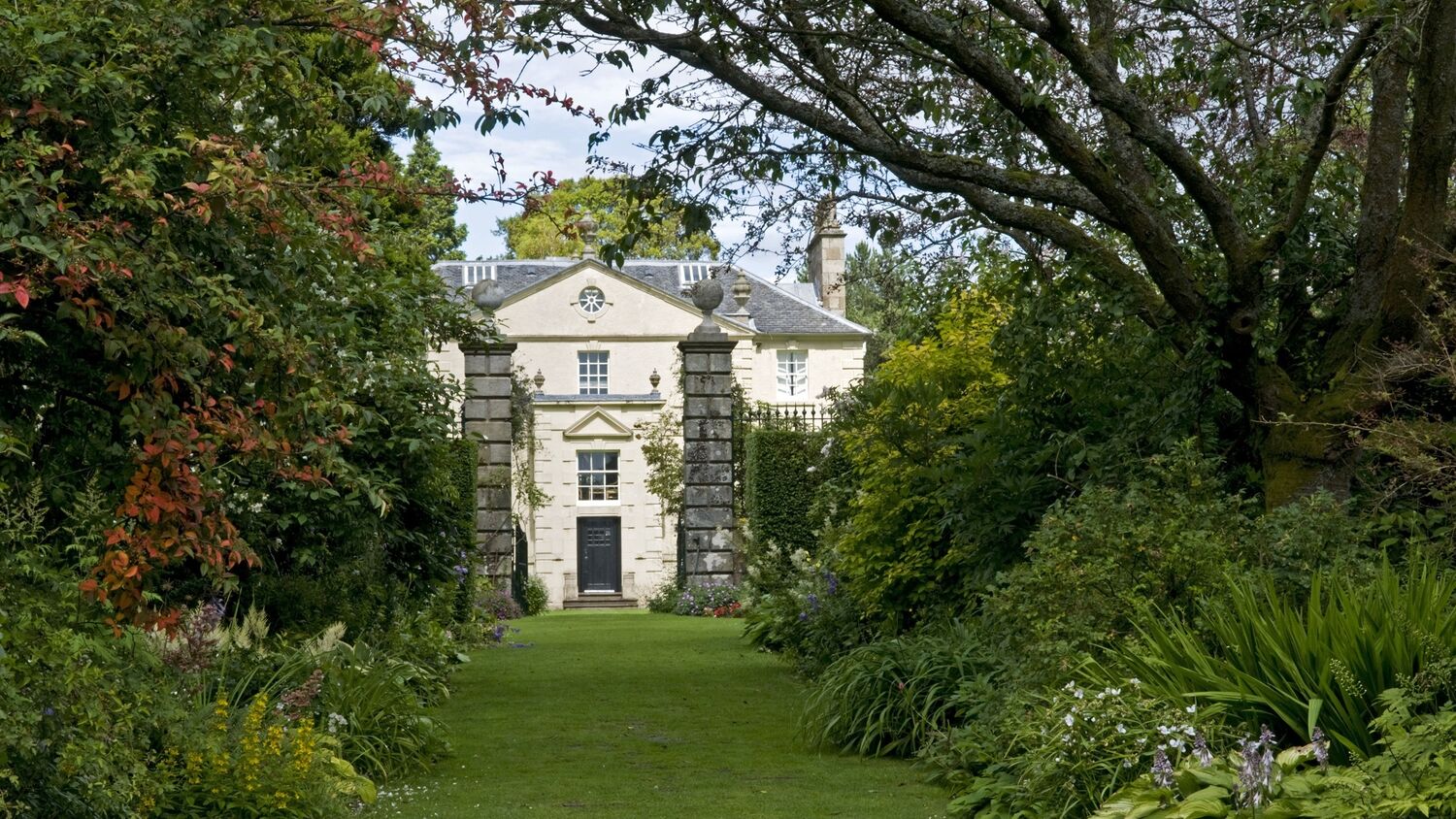 A view of the white stone 18th-century Greenbank House, seen from between two stone pillars in the garden. Lush flower beds run down either side from the gate pillars, filled with colourful plants, shrubs and trees.