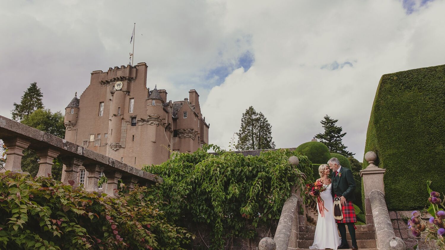 A bride and groom stand on stone garden steps, with Crathes Castle in the background. The groom wears a red kilt. The bride wears a long white dress and is carrying a beautiful bouquet of red and orange flowers.