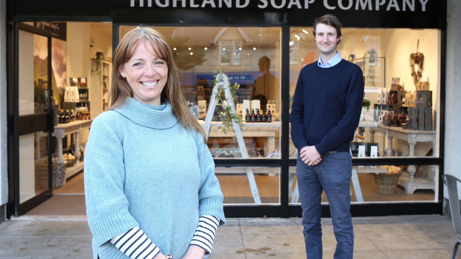 A woman and a man pose smilingly in front of a shop entrance and store window, that has the sign above it reading Highland Soap Company.